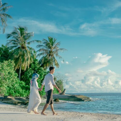 A couple holding hands while walking on a serene tropical beach with palm trees and clear skies.