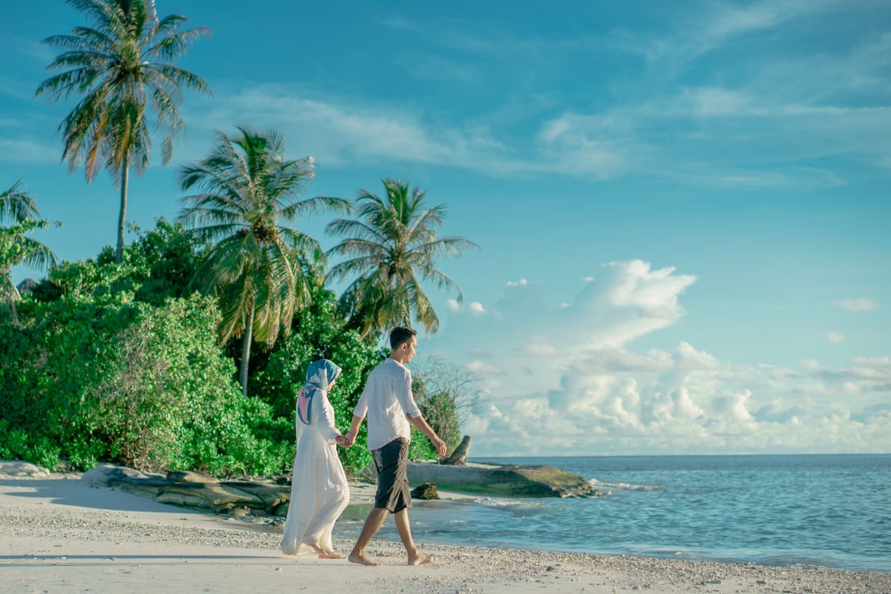 A couple holding hands while walking on a serene tropical beach with palm trees and clear skies.